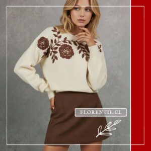 Modelo rubia de frente con sweater bordado flores crema y rosas chocolate, falda mini marrón, mano cerca del mentón, fondo gris claro