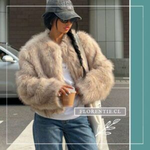 Modelo con chaqueta abrigo efecto pelo beige camel, gorra, camiseta blanca visible y jeans, sosteniendo café en exterior urbano.