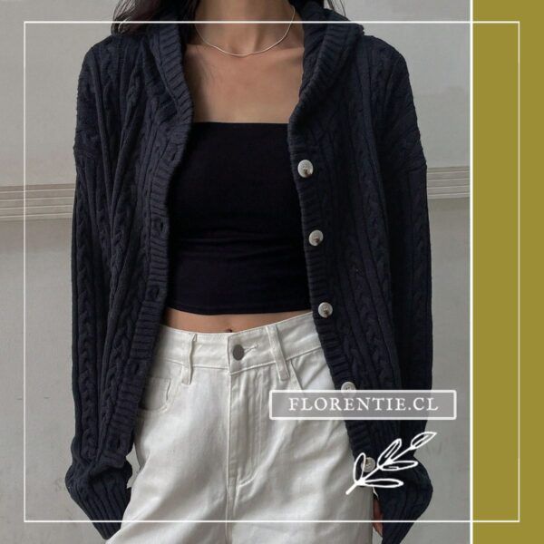 Modelo con cardigan azul marino abotonado, capucha y trenzas cable knit, manos en bolsillos, combinado con pantalón blanco, look exterior.