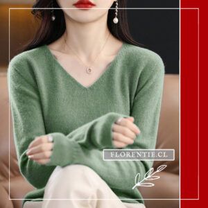 Modelo sentada en sofá luciendo sweater verde oliva de lana merino 100% con escote V y mangas largas con puños acanalados sobre falda blanca.
