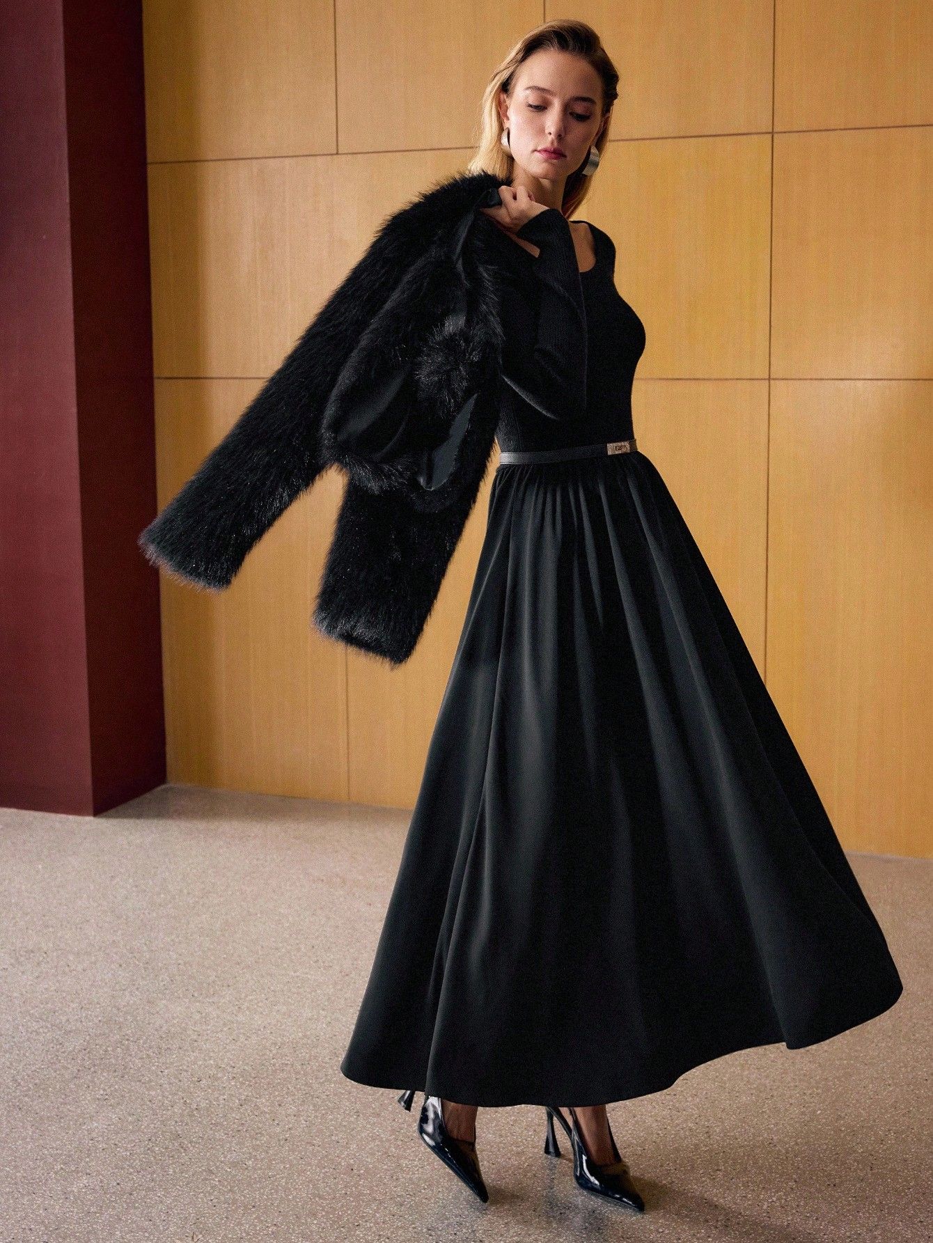 Modelo luciendo vestido negro largo con falda amplia y chaqueta negra de pelo sobre los hombros, tacos charol negros, look elegante de otoño