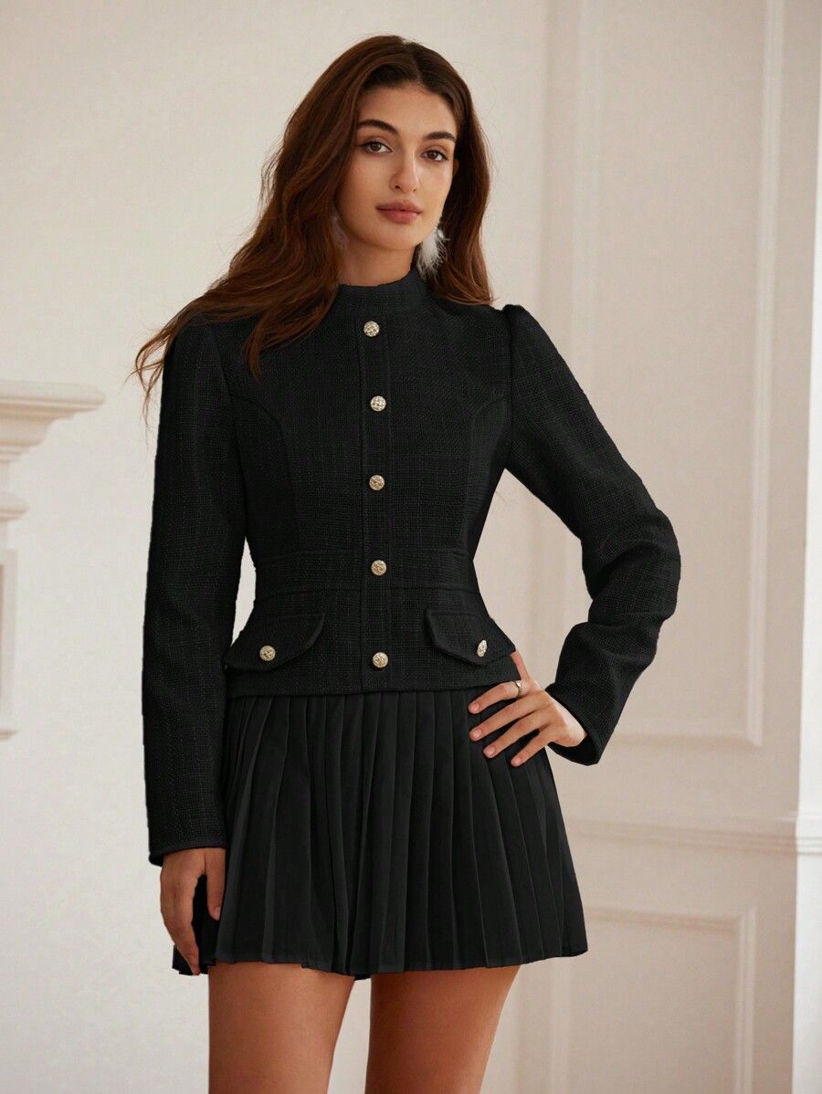 Mujer con pose elegante vistiendo vestido blazer negro entallado con cuello alto y falda plisada mano en la cintura fondo clásico