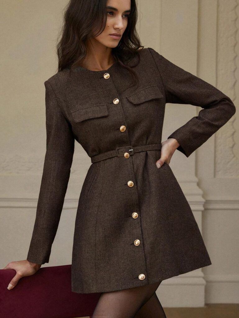Modelo posando con vestido blazer tweed espiga café, botones dorados frontales, cinturón en cintura y bolsillos con solapa sobre fondo beige elegante