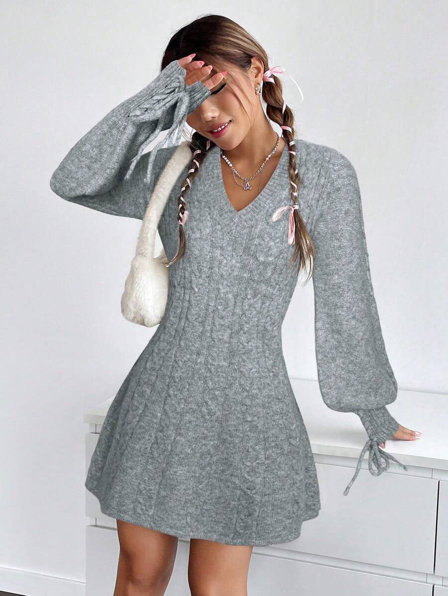 Modelo posando con vestido gris de punto trenzado, escote en V, mangas globo con lazo en puños, falda acampanada y bolso blanco de peluche