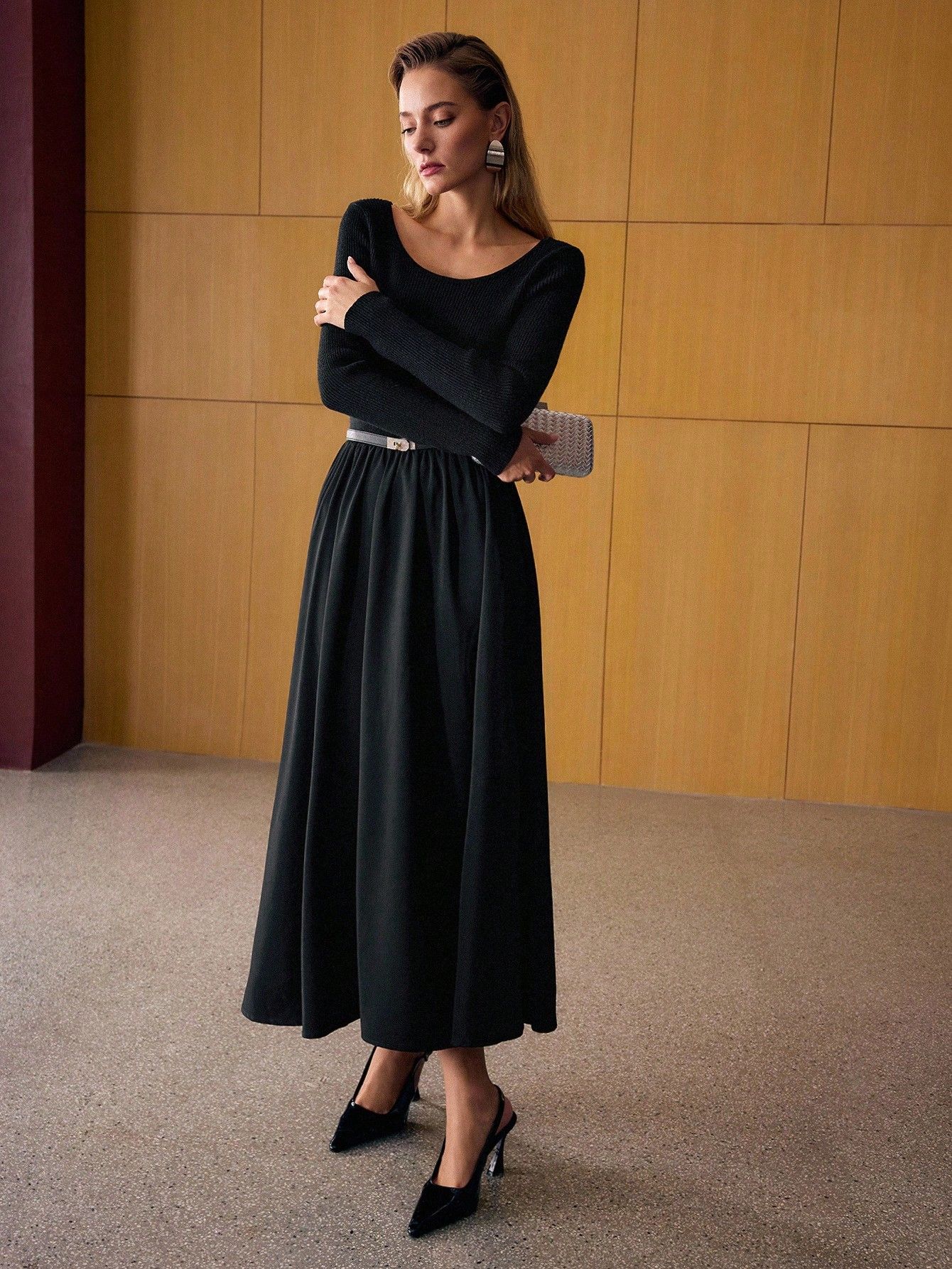 Modelo en pose lateral con vestido negro de cuerpo entallado acanalado y falda maxi amplia, clutch beige tejido, fondo paneles madera dorada
