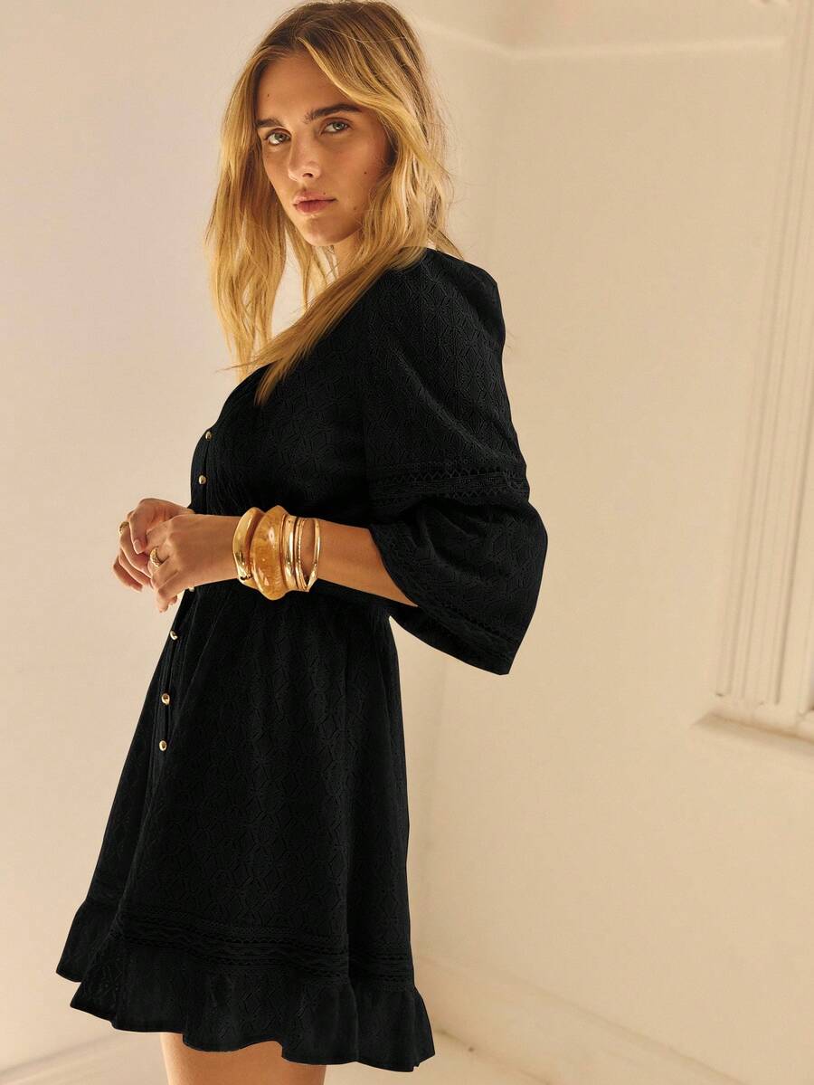 Modelo posando de perfil con vestido negro de escote en V, falda acampanada con volante y manga larga texturizada, brazaletes dorados como accesorios