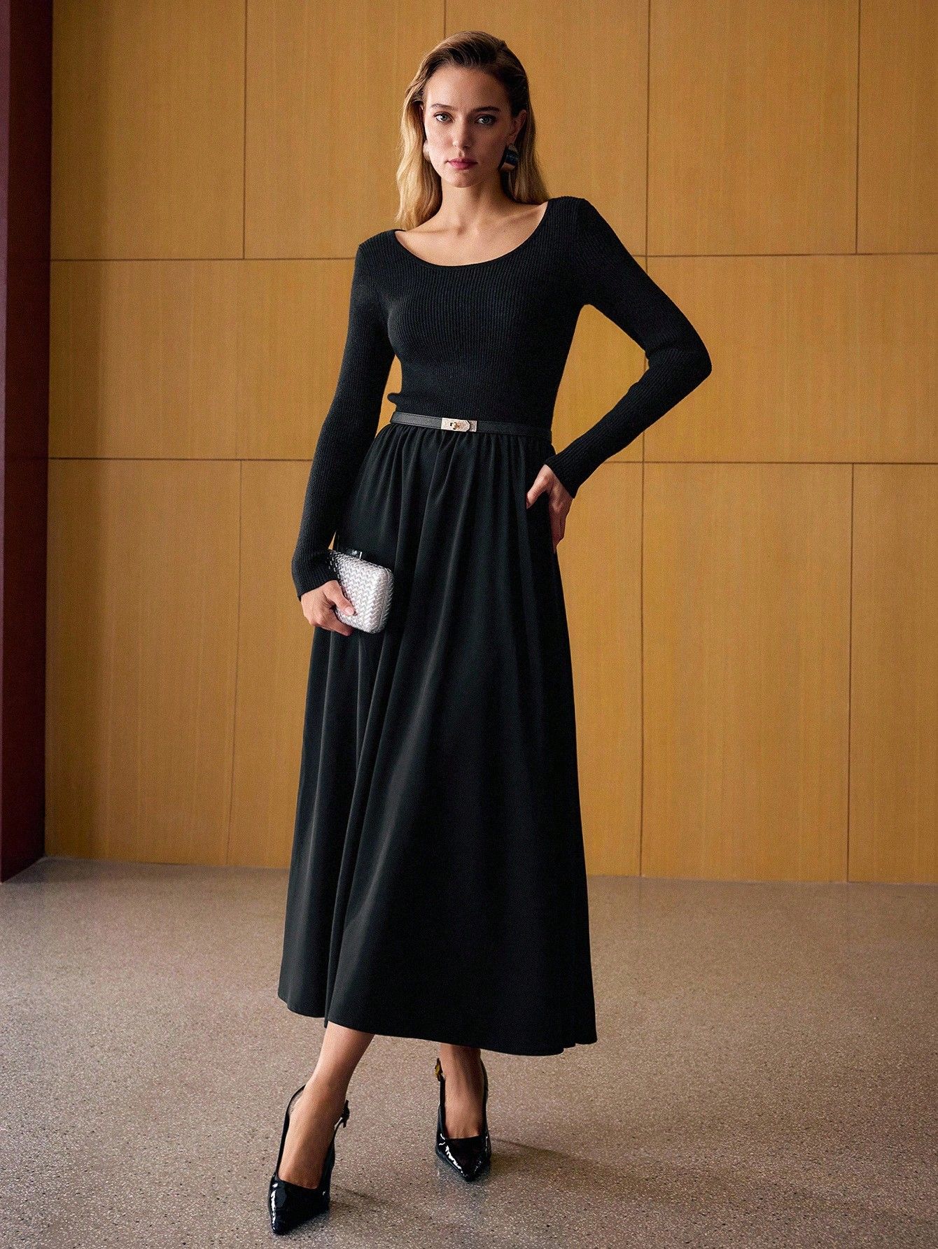 Mujer de frente con vestido negro largo manga larga, falda amplia fluida, cinturón fino, sosteniendo clutch plateado tejido, tacos negros charol