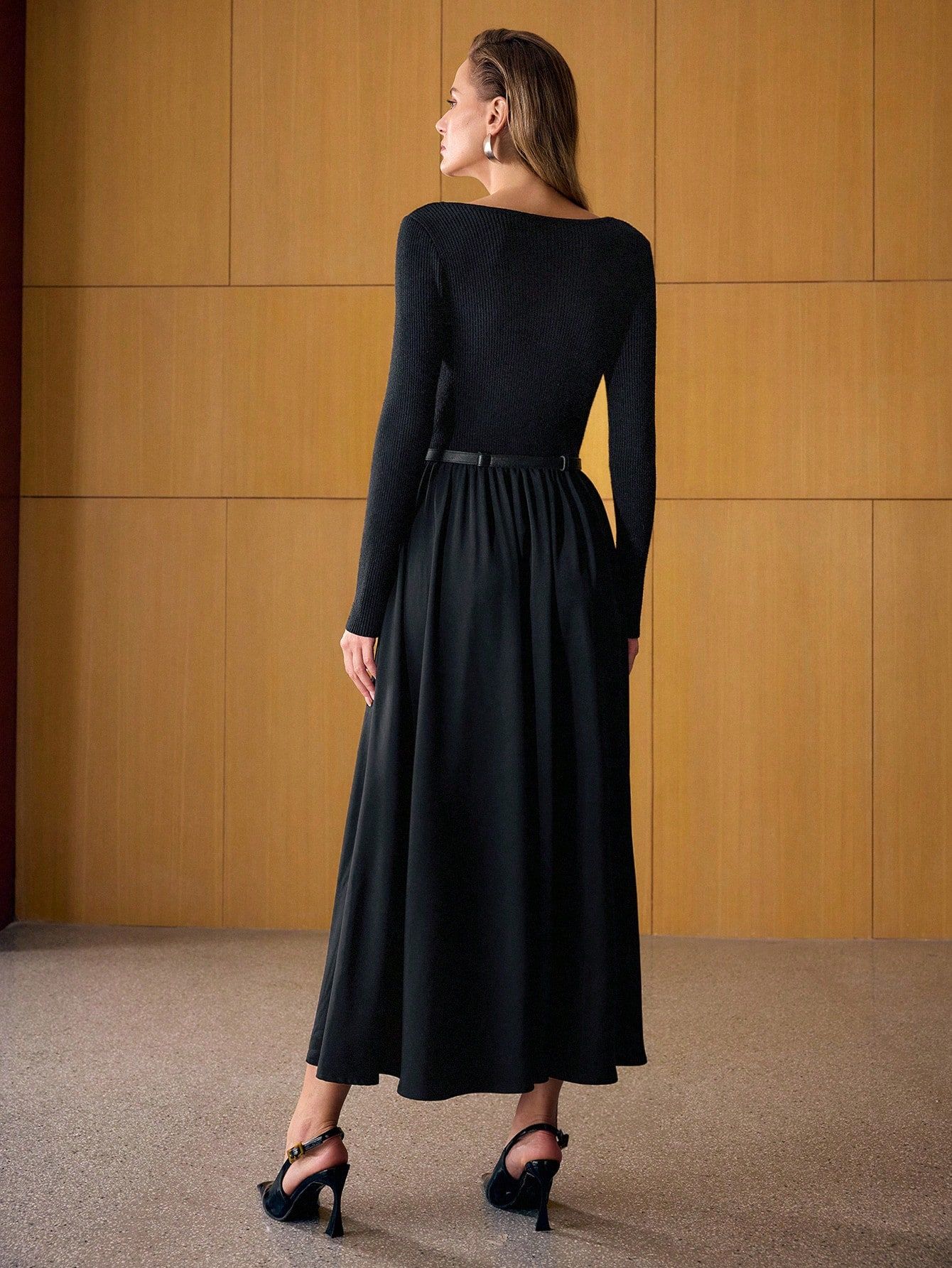 Vista posterior de modelo con vestido negro largo, espalda tejido acanalado, falda amplia con caída elegante, cinturón fino, tacos negros con detalle dorado