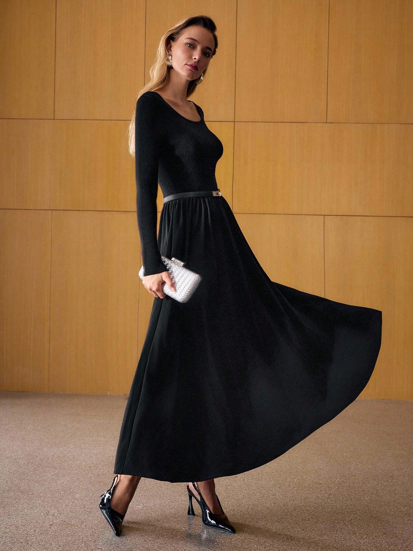 Mujer caminando con vestido negro largo mostrando el movimiento de la falda amplia fluida, manga larga tejida, clutch plateado, tacos charol negros