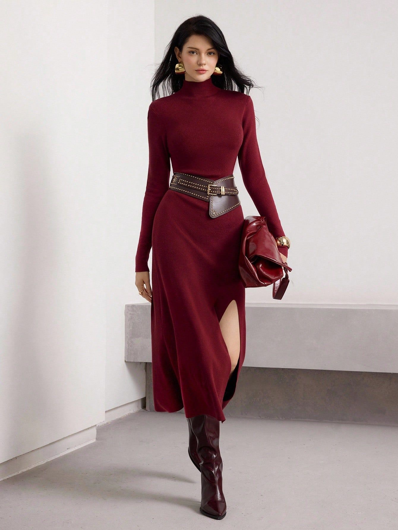 Modelo con vestido de punto burdeos cuello alto y manga larga, cinturón marrón con tachas, cartera roja y botas altas en fondo minimalista