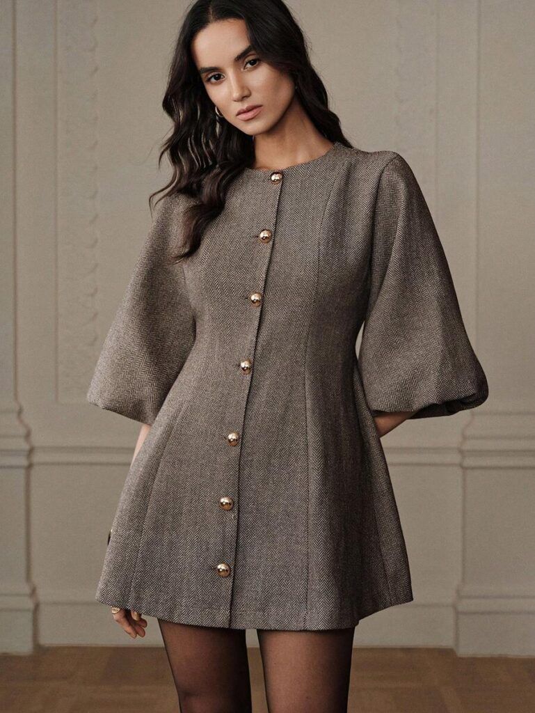 Modelo con vestido corto de tweed espigado café con botones dorados, manga campana tres cuartos, pose frontal elegante con fondo clásico