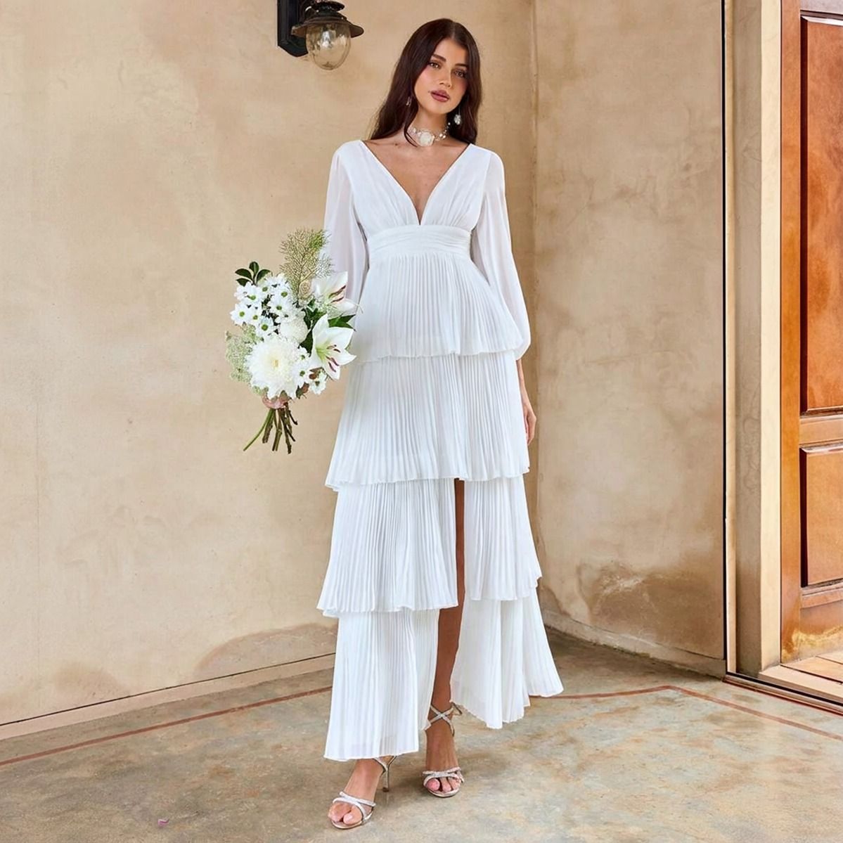 Vestido blanco maxi con cuatro capas plisadas, escote V profundo y mangas globo, vista frontal con ramo de flores