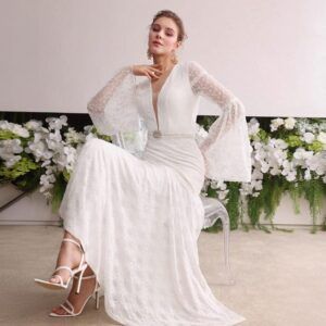 Modelo sentada con vestido blanco de encaje largo, escote V profundo y mangas campana transparentes con cinturón de perlas