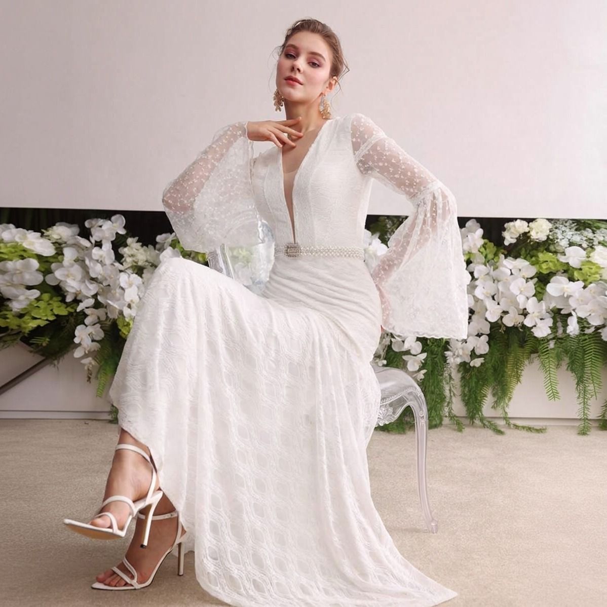 Modelo sentada con vestido blanco de encaje largo, escote V profundo y mangas campana transparentes con cinturón de perlas