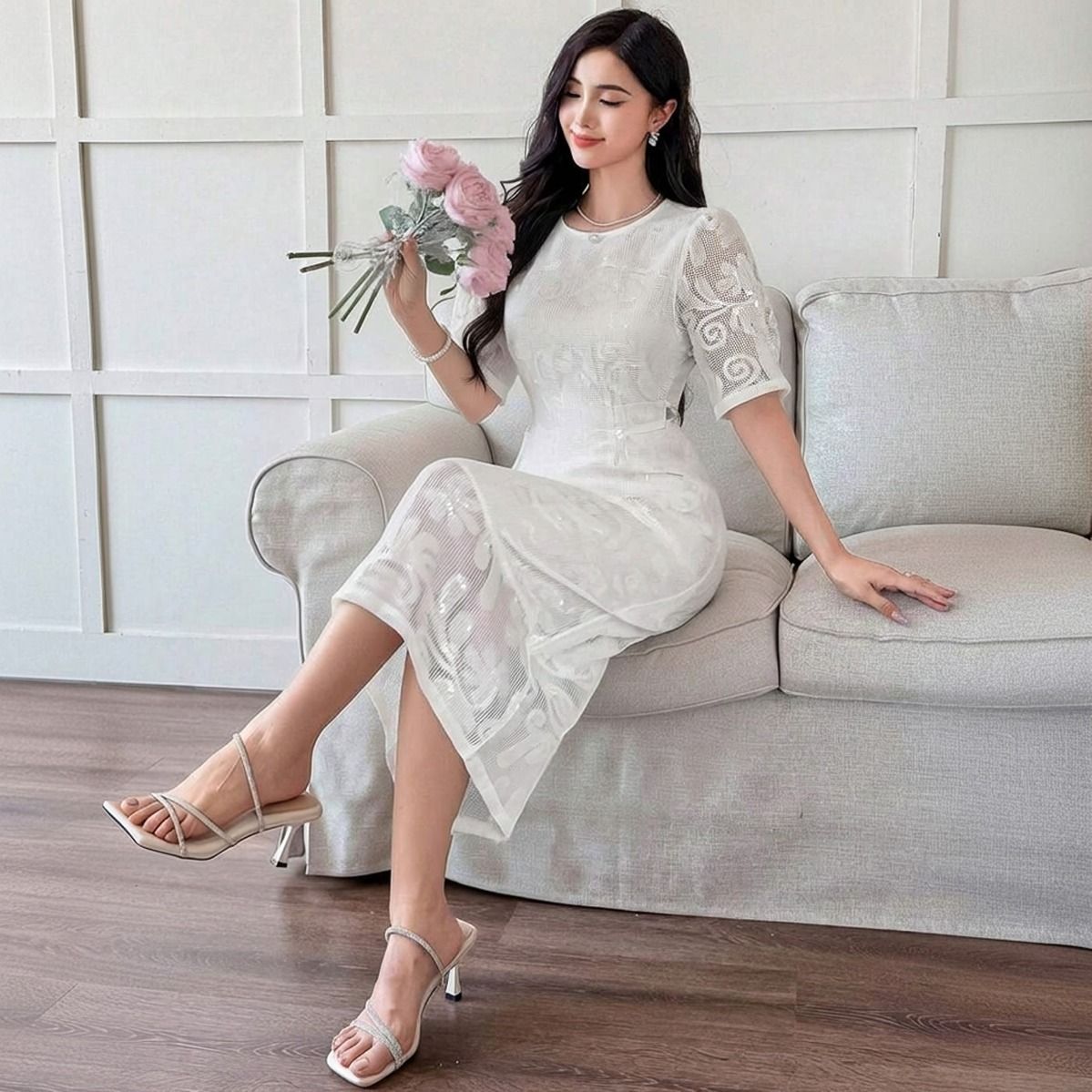 Modelo sentada en sofá sosteniendo ramo de flores con vestido midi blanco de encaje y manga corta transparente