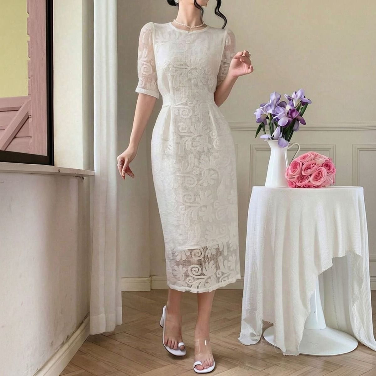 Vestido midi blanco de encaje calado con forro interior, de pie junto a mesa con flores, vista frontal completa