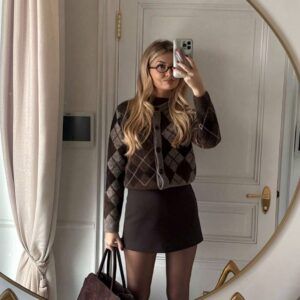 Mujer con cardigan argyle café y gris cropped combinado con minifalda marrón y medias opacas frente a espejo
