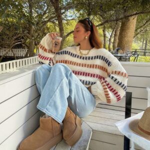 Mujer sentada en banco blanco con sweater crudo de rayas multicolor manga ancha y jeans claros