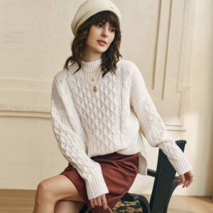 Sweater blanco tejido con trenzas y cuello mock neck, modelo sentada vista frontal