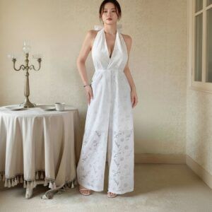 Mujer de pie con enterito blanco de broderie, cuello halter y pierna palazzo, junto a mesa con candelabro