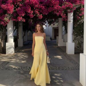 Mujer de pie usando vestido maxi amarillo pastel de lino con tirantes finos y falda evasé bajo un arco de flores
