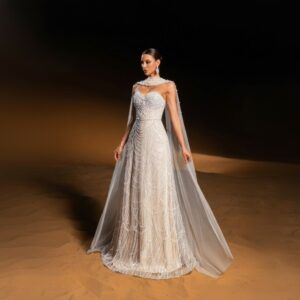 Vestido largo marfil bordado en perlas con capa de tul, vista frontal de cuerpo completo sobre arena