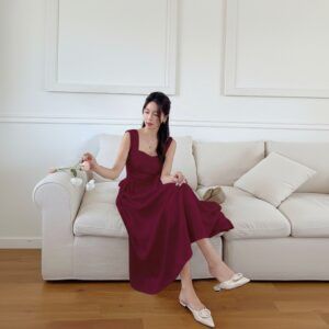 Mujer sentada en sofá con vestido midi burdeos de escote corazón y tirantes anchos, falda evasé sobre las rodillas