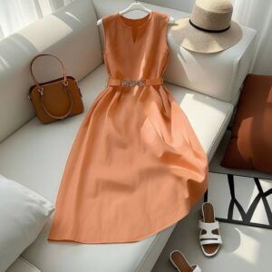 Vestido midi color melocotón sin mangas con escote notch y cinturón de cadena dorada, tendido sobre sofá blanco