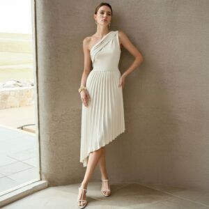 Vestido midi crema con escote un hombro drapeado y falda plisada asimétrica, vista frontal apoyada en pared