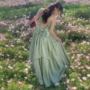Mujer de espaldas con vestido largo verde salvia en satén con lazo grande en la espalda descubierta, en campo de flores